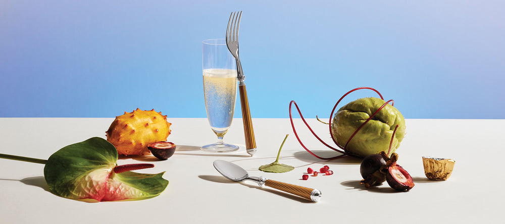 Champagne Flute and Cable Silverware arranged on a tabe with tropical fruits and a light blue background