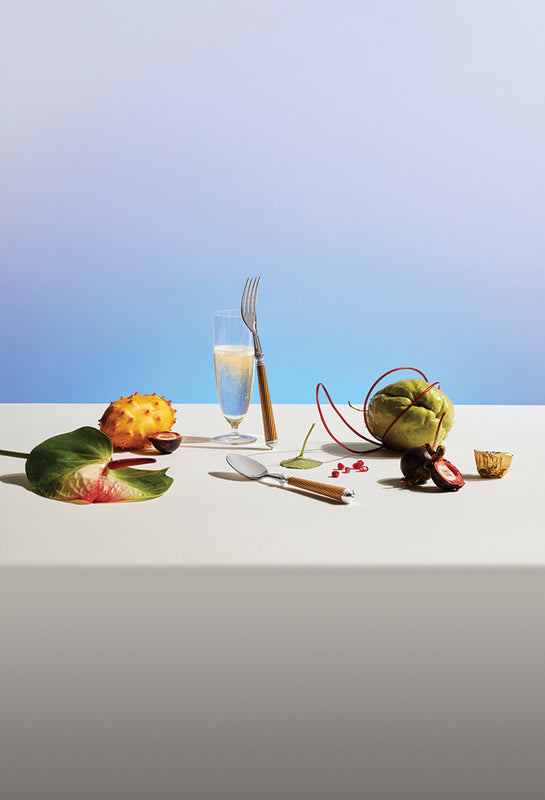 Champagne Flute and Cable Silverware arranged on a tabe with tropical fruits and a light blue background