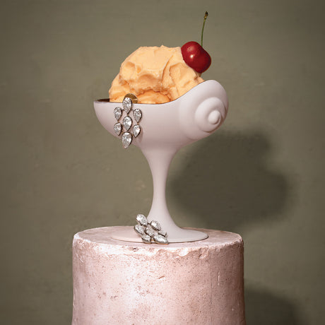 A sculptural white dessert coupe bowl filled with orange ice cream and topped with a red cherry, displayed on a pale pink pedestal against a muted olive-gray background. Silver jeweled floral accents are attached to the bowl and stem.