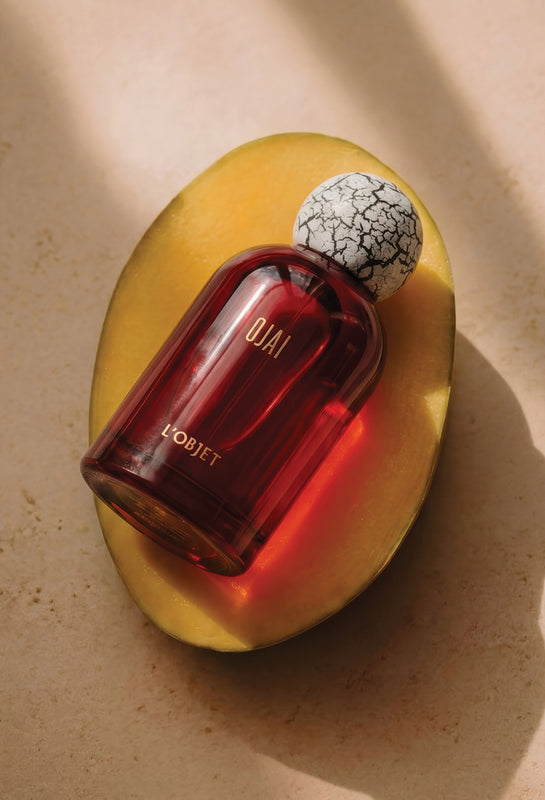 A YORJET perfume bottle with a deep amber-red glass body and a white-and-black crackled cap, resting on a sliced yellow mango against a warm beige background.