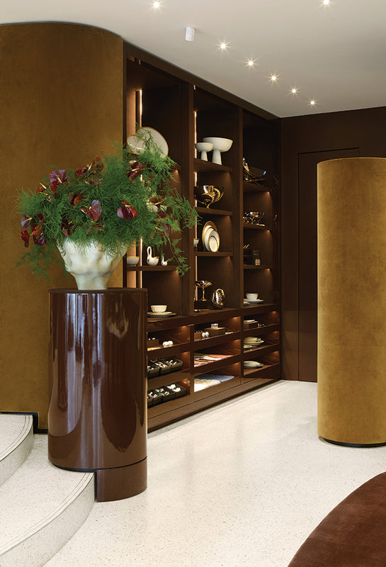 A luxury interior design scene featuring a dark brown glossy pedestal table holding a white vase with green foliage and red flowers, set against a background of built-in wooden shelves with decorative items, a gold velvet cylinder column, and a white terrazzo floor.