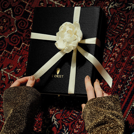 A black gift box wrapped with a cream ribbon and a white fabric flower, held by two hands wearing sparkly brown sleeves. The box has the brand name 'L'OBJET' printed in gold on the front, set against a dark red patterned rug.