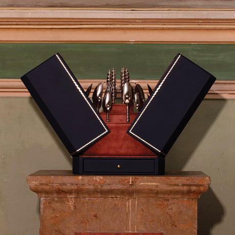 A decorative black presentation box with open wings, lined with red velvet and trimmed with white stitching, holding a set of ornate silver horse-shaped utensils or tools, displayed on a stone pedestal against an indoor wall.