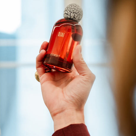 A hand holding a bottle of Eau de Parfum, showcasing the translucent amber-red glass and the distinctive cracked black-and-white ceramic cap, set against a softly blurred indoor background.
