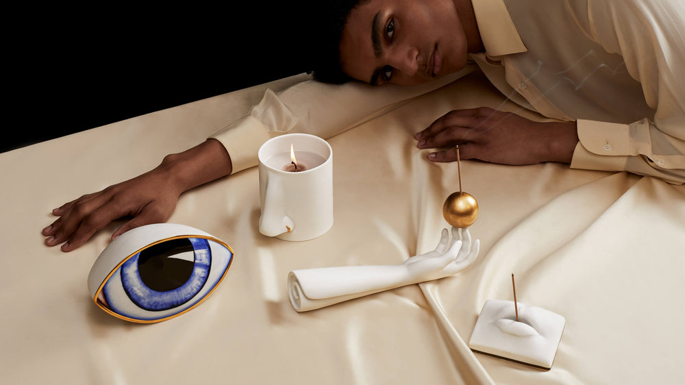 Person lying on a bed with decorative items including a candle, eye-shaped object, and gold sphere.
