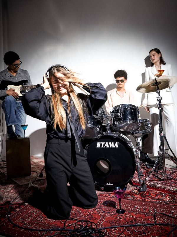 A band performing live on stage, with a woman in the foreground wearing headphones and smiling, a man playing guitar on the left, a drummer behind her, and a woman holding a drink on the right. A TAMA bass drum is visible in the center.