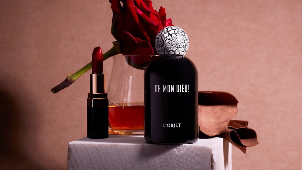 Discover Eau de Parfum. Shop L'OBJET Fragrance Collection. Oh Mon Dieu! Black Porcelain Fragrance Bottle with Cracked Wooden Cap. On Stone pedastal with cinnamon, whiskey, rose and lipstick around it.