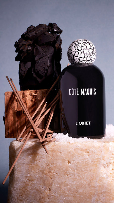 Shop Cote Maquis and other scents from our fragrance collection. Black porcelain bottle with white crackled top on on beige stone with cinnamon, incense and black essence in front of a shadow cast light blue grey background.