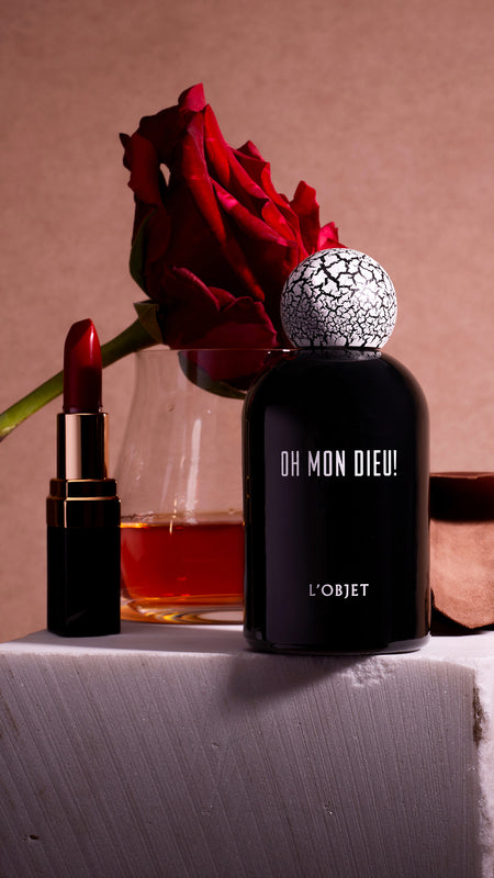 Discover Eau de Parfum. Shop L'OBJET Fragrance Collection. Oh Mon Dieu! Black Porcelain Fragrance Bottle with Cracked Wooden Cap. On Stone pedastal with cinnamon, whiskey, rose and lipstick around it.