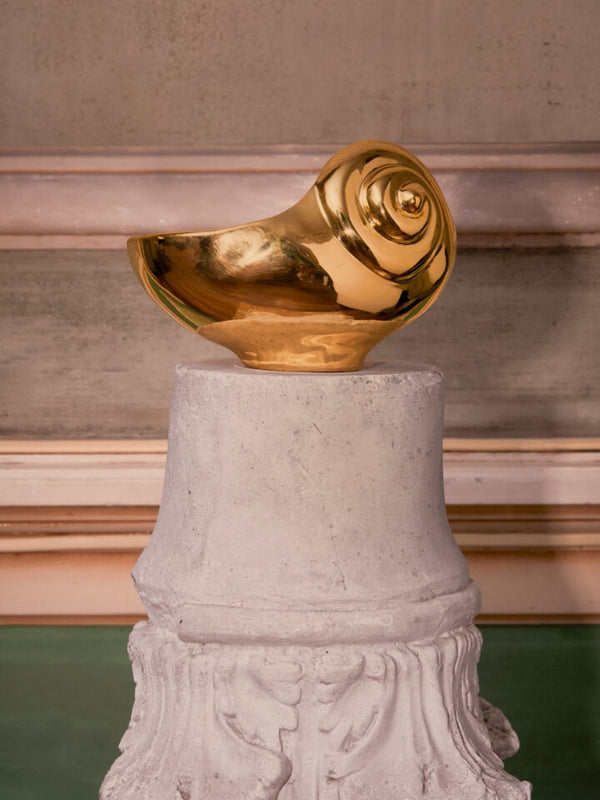 A polished gold shell-shaped sculpture or decorative object displayed on top of a white stone pedestal against an indoor architectural background.